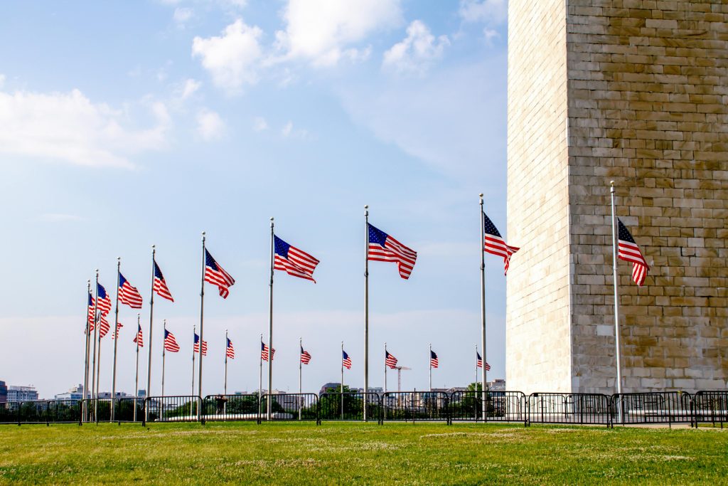 American flags flying at the base of the Washington Monument against a blue sky.